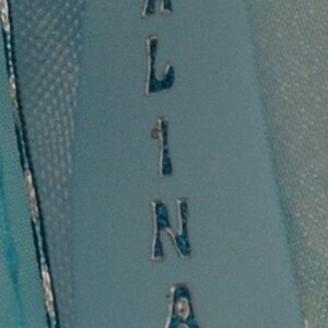 Puede incluir: Una cinta azul claro con el nombre "ALINA" escrito en letras plateadas con detalles de purpurina azul.