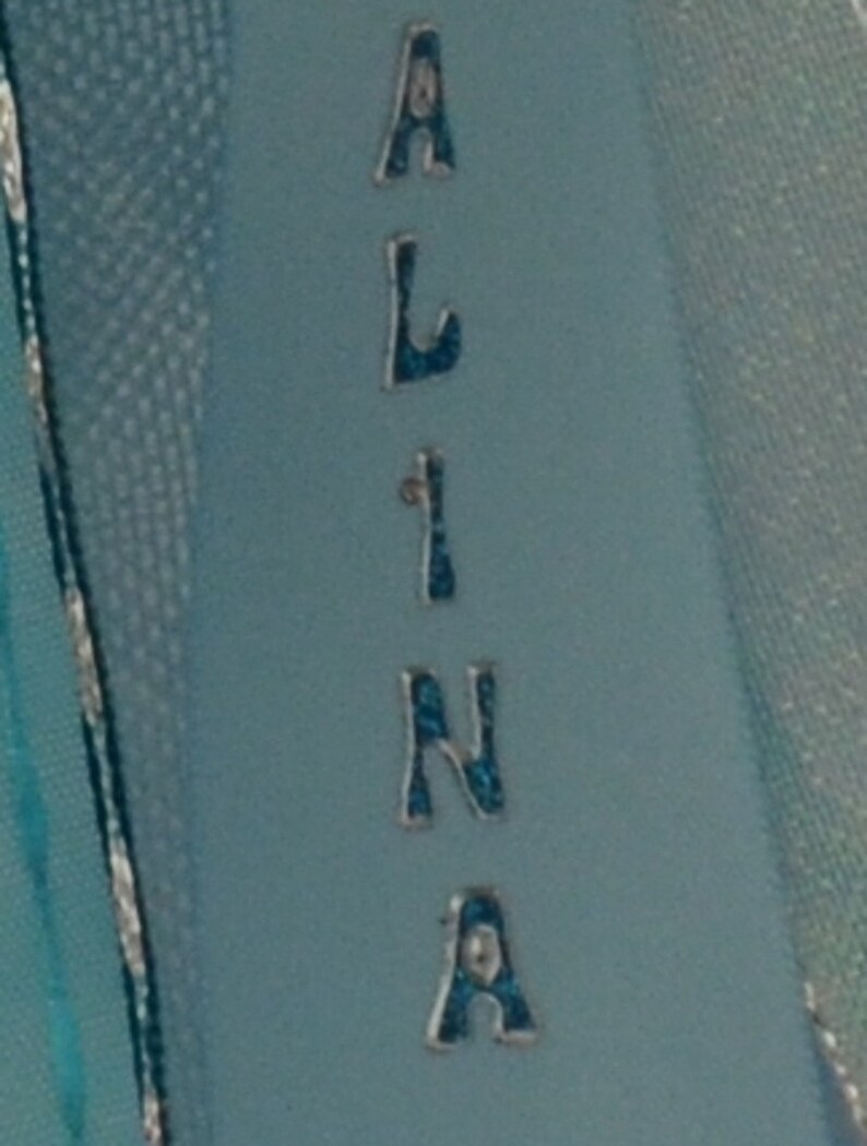 Puede incluir: Una cinta azul claro con el nombre "ALINA" escrito en letras plateadas con detalles de purpurina azul.
