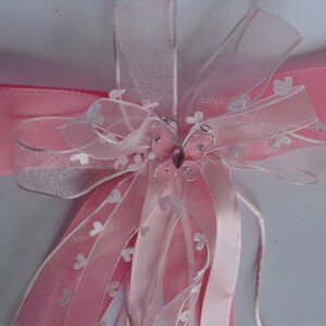 Puede incluir: Lazo de cinta de tul rosa y blanco con un adorno de mariposa plateada. La mariposa tiene una gema rosa en el centro.