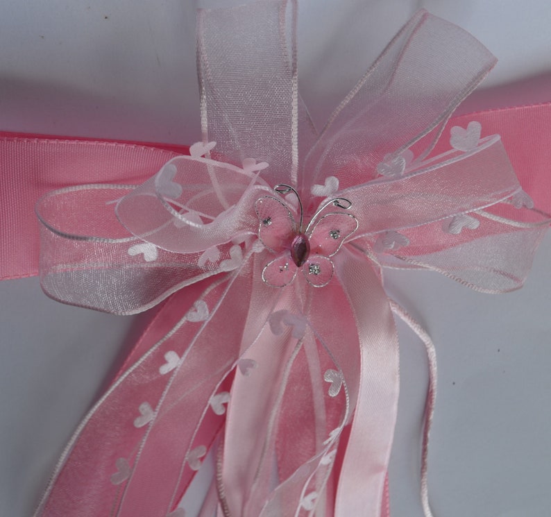 Puede incluir: Lazo de cinta de tul rosa y blanco con un adorno de mariposa plateada. La mariposa tiene una gema rosa en el centro.