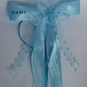 Puede incluir: Una cinta azul claro con un lazo y un peque&ntilde;o amuleto de mariposa. La cinta tiene la palabra "NAME" impresa.