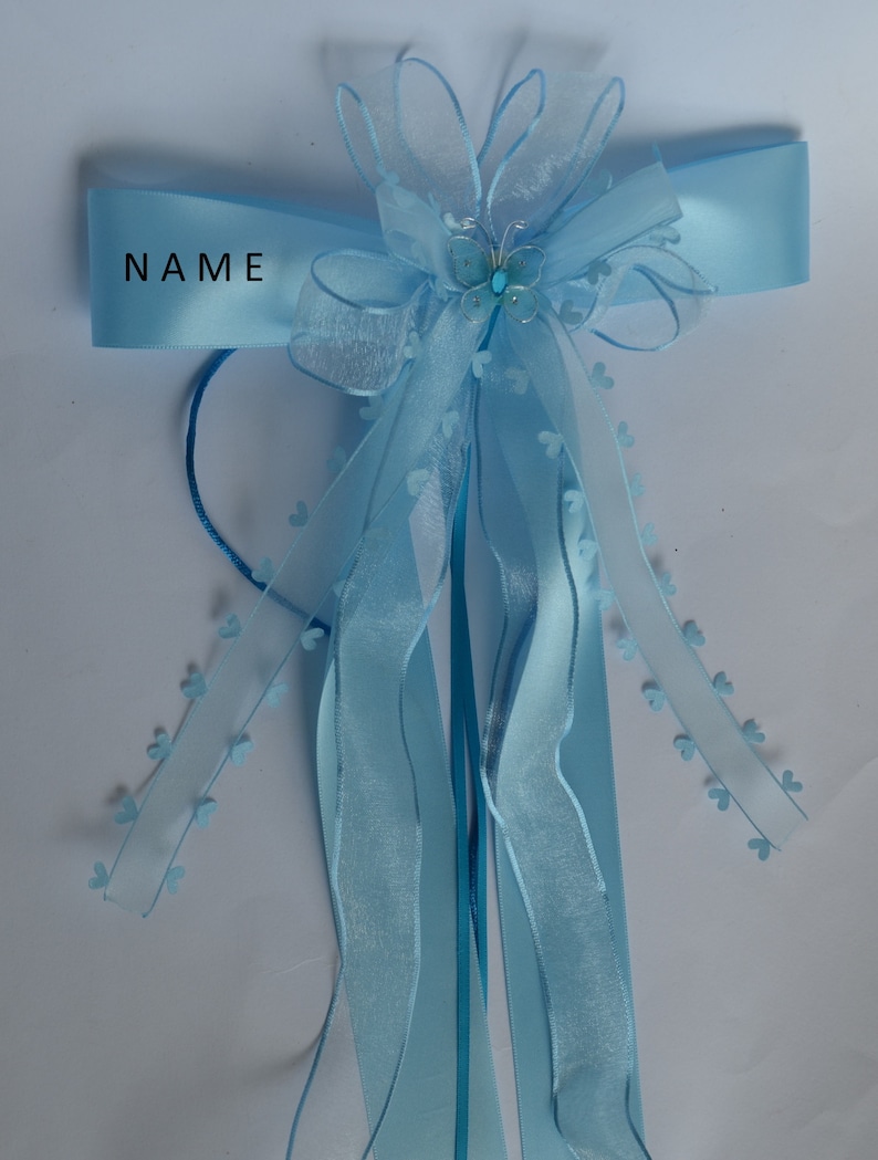 Puede incluir: Una cinta azul claro con un lazo y un peque&ntilde;o amuleto de mariposa. La cinta tiene la palabra "NAME" impresa.