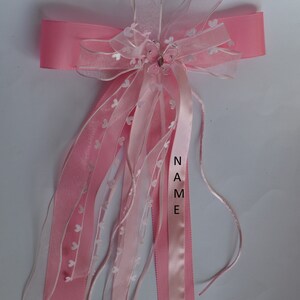 Puede incluir: Lazo de cinta rosa y blanco con un dije de mariposa y la palabra "NAME" escrita en la cinta. El lazo est&aacute; decorado con peque&ntilde;os corazones blancos.