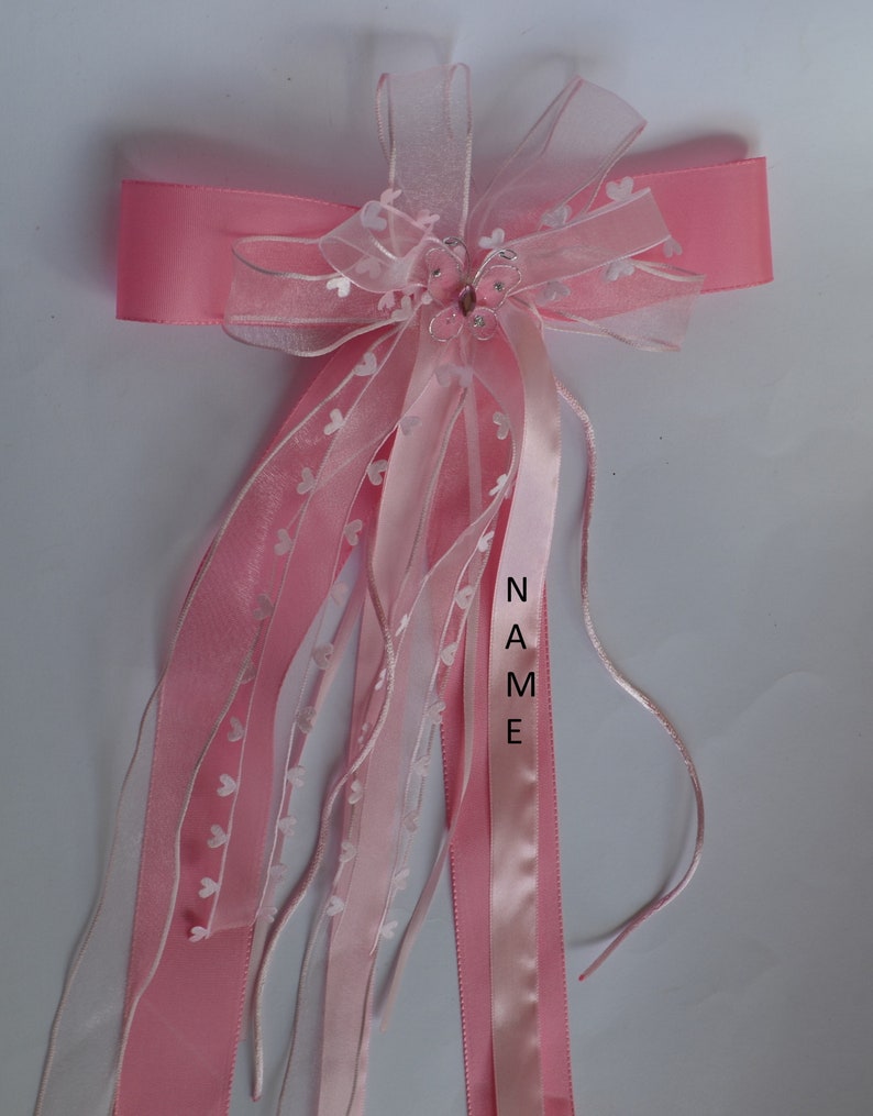 Puede incluir: Lazo de cinta rosa y blanco con un dije de mariposa y la palabra "NAME" escrita en la cinta. El lazo est&aacute; decorado con peque&ntilde;os corazones blancos.