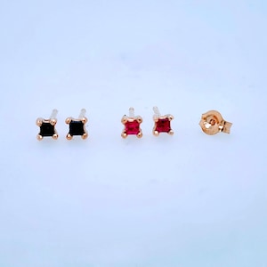 May include: Two pairs of square-cut stud earrings with gold-toned settings. One pair features deep black stones, while the other pair features vibrant red stones. A single gold-toned earring back is positioned to the right of the red earrings.