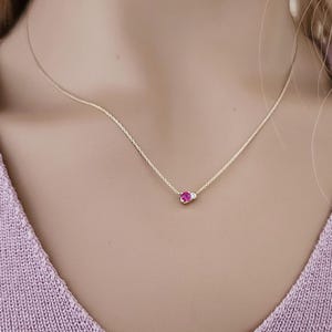 May include: A delicate gold chain necklace featuring a small, round, vibrant pink gemstone set next to a tiny, clear, round white gemstone. The necklace rests against a light purple knit fabric, showcasing a minimalist design with a thin metal chain.