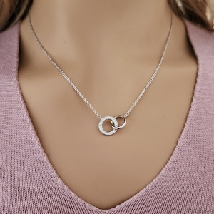 May include: A silver necklace featuring two interlocking circles. One circle is encrusted with small, sparkling stones, while the other is a plain silver band. The necklace hangs on a delicate chain, creating a simple yet elegant design.