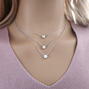 May include: A layered necklace set with three delicate chains. Each chain features a different shaped, clear gemstone: a rectangle, a square, and a circle. The necklaces are silver in color and are displayed on a person wearing a pink sweater.