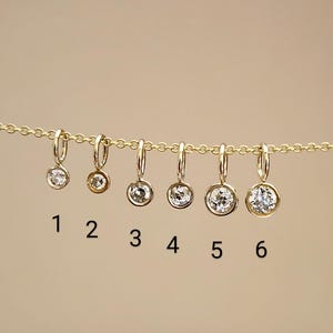 May include: Six gold-colored hoop earrings, each with a clear, round gemstone. The earrings are numbered 1 through 6. The earrings are suspended from a gold chain against a neutral background.