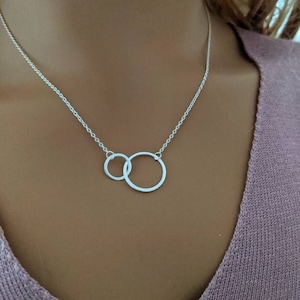 May include: A silver necklace featuring two interlocking circles. The circles are connected by a delicate chain, creating a minimalist and elegant design. The necklace is shown against a neutral background.