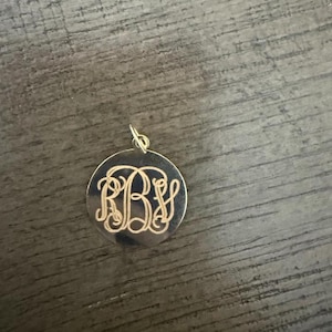 Puede incluir: Un colgante circular de tono dorado que presenta un monograma grabado intrincado con las letras R, B y S entrelazadas en un estilo de escritura decorativo. El colgante incluye una peque&ntilde;a anilla de metal en la parte superior.