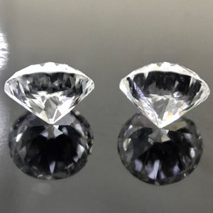 May include: Two clear, round, faceted gemstones, likely diamonds, are shown on a dark gray surface. The gemstones are reflecting light and casting shadows.