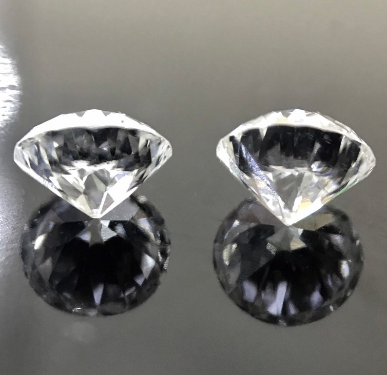 May include: Two clear, round, faceted gemstones, likely diamonds, are shown on a dark gray surface. The gemstones are reflecting light and casting shadows.