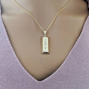 Op de afbeelding: Een goudkleurige ketting met een rechthoekige hanger met het woord "GOLD" in reli&euml;f. De hanger hangt aan een fijne gouden ketting. De ketting wordt getoond tegen een zachte, lichtpaarse achtergrond.