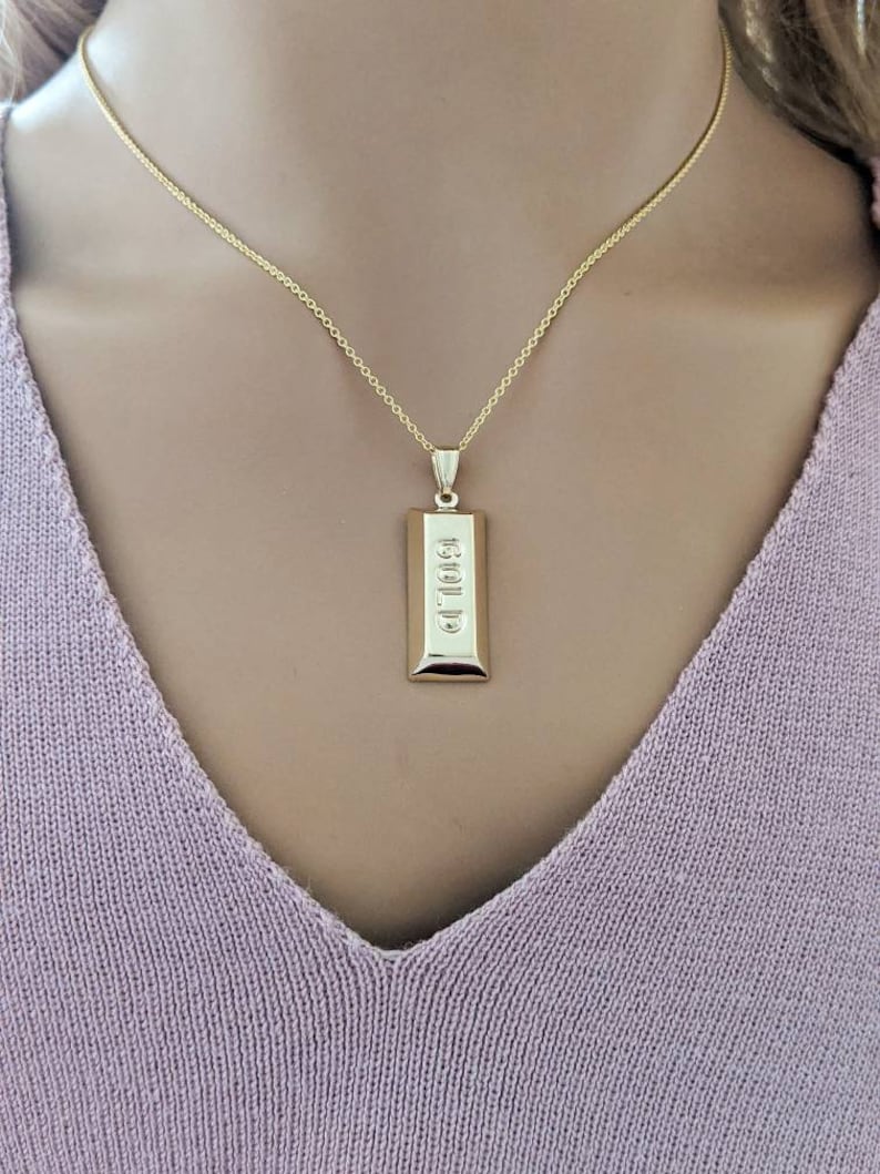 Op de afbeelding: Een goudkleurige ketting met een rechthoekige hanger met het woord "GOLD" in reli&euml;f. De hanger hangt aan een fijne gouden ketting. De ketting wordt getoond tegen een zachte, lichtpaarse achtergrond.