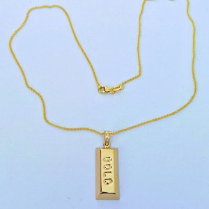 Op de afbeelding: Een goudkleurige ketting met een rechthoekige hanger met het woord "GOLD" gegraveerd. De hanger hangt aan een fijne gouden ketting met een karabijnsluiting.