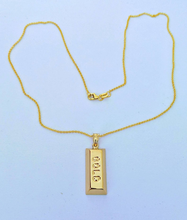 Op de afbeelding: Een goudkleurige ketting met een rechthoekige hanger met het woord "GOLD" gegraveerd. De hanger hangt aan een fijne gouden ketting met een karabijnsluiting.