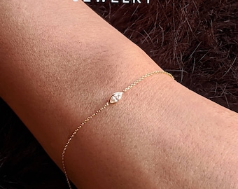 May include: A delicate gold chain bracelet with a small diamond-shaped pendant. The pendant is shaped like an eye and is set in a gold setting. The bracelet is shown on a person's wrist.