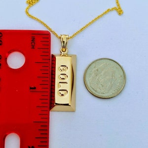 Op de afbeelding: Een goudkleurige hangerketting met een rechthoekig staafontwerp met het woord "GOLD" in reli&euml;f. De hanger is ongeveer 2,5 cm lang. Een gouden ketting en een munt zijn ook zichtbaar.