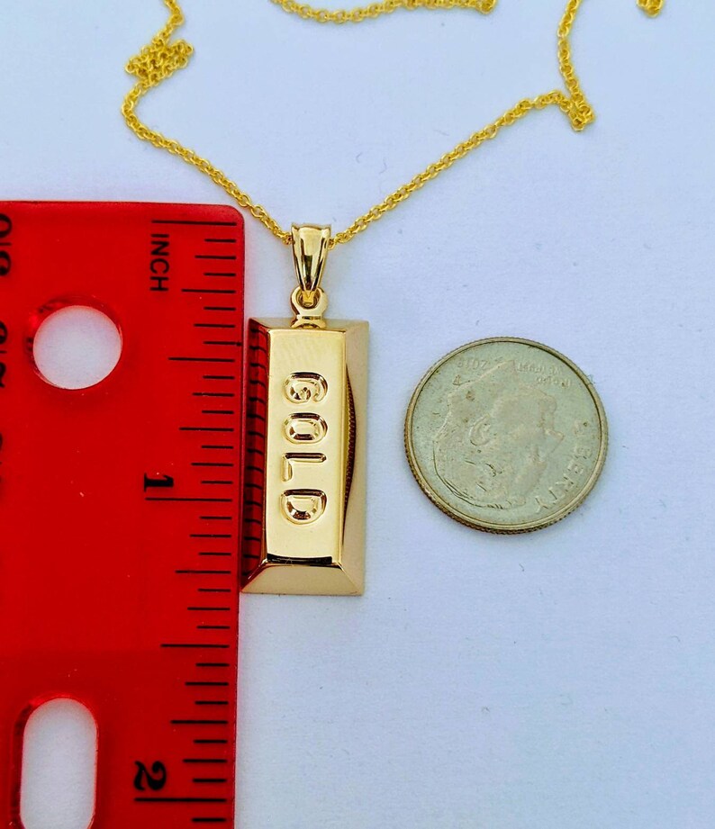 Op de afbeelding: Een goudkleurige hangerketting met een rechthoekig staafontwerp met het woord "GOLD" in reli&euml;f. De hanger is ongeveer 2,5 cm lang. Een gouden ketting en een munt zijn ook zichtbaar.