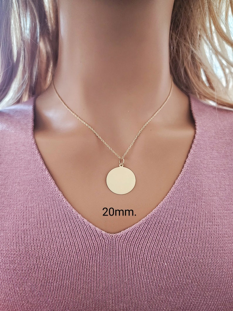 Puede incluir: Un collar de color dorado con un colgante circular de superficie lisa y plana. El colgante cuelga de una cadena delicada. El texto 20mm. es visible debajo del colgante, indicando el di&aacute;metro del dije circular.