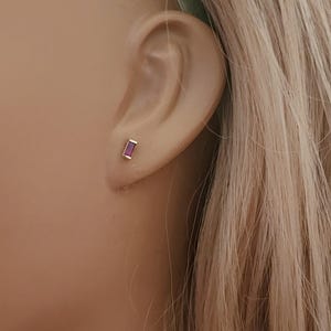 May include: A small gold stud earring featuring a rectangular baguette-cut purple gemstone. The earring is positioned in the earlobe, showcasing a minimalist metal setting that secures the vibrant, deep purple stone in a vertical orientation.