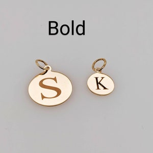 Puede incluir: Dos dijes redondos de metal en tono dorado con anillas, cada uno con una letra grabada. El dije m&aacute;s grande muestra la letra S y el m&aacute;s peque&ntilde;o la letra K. La palabra Bold est&aacute; impresa en texto negro sobre los dijes.