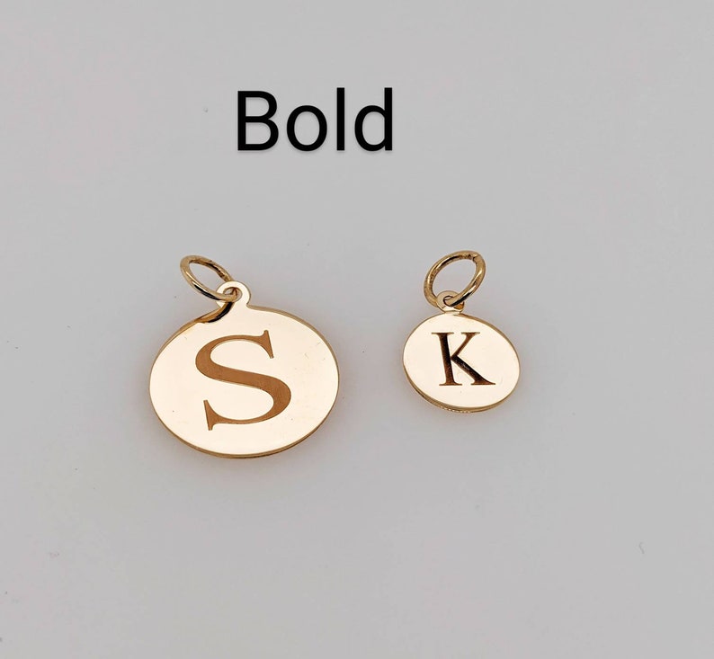 Puede incluir: Dos dijes redondos de metal en tono dorado con anillas, cada uno con una letra grabada. El dije m&aacute;s grande muestra la letra S y el m&aacute;s peque&ntilde;o la letra K. La palabra Bold est&aacute; impresa en texto negro sobre los dijes.