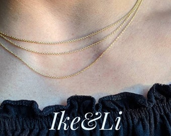 May include: Three gold chain necklaces layered on a person's neck. The necklaces are delicate and have a subtle shine. The image is cropped at the neck and shoulders, with a black shirt visible below. The text "Ike & Li JEWELRY" is visible at the bottom of the image.