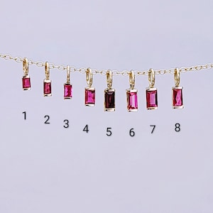 May include: A gold chain necklace featuring eight rectangular baguette-cut pink gemstone charms hanging from jump rings. Each charm is numbered one through eight in black text below it, showcasing variations in size and color intensity.