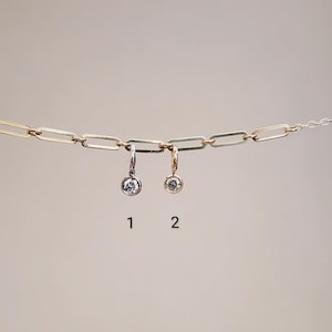 May include: Two delicate earrings with a round, clear gemstone are suspended from a gold chain. One earring is silver-toned, the other gold-toned. The background is a soft, neutral color.
