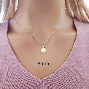 Puede incluir: Un collar de color dorado con una cadena fina y un peque&ntilde;o colgante circular. El colgante tiene un di&aacute;metro de 8 mm. El collar descansa sobre una tela texturizada de color rosa claro.