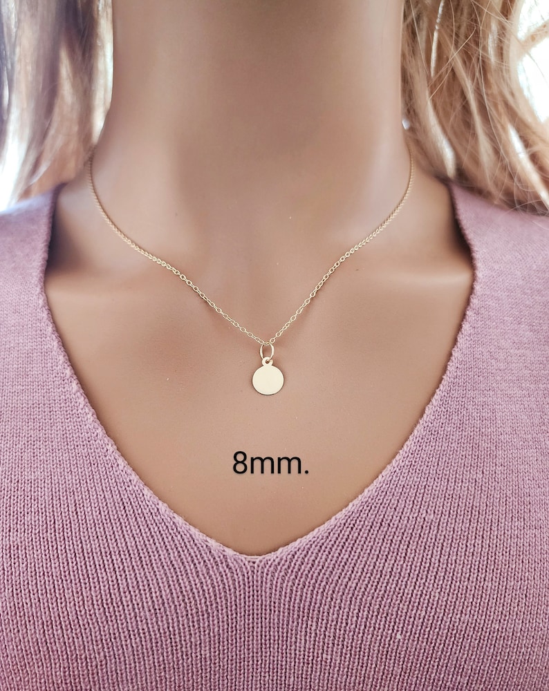 Puede incluir: Un collar de color dorado con una cadena fina y un peque&ntilde;o colgante circular. El colgante tiene un di&aacute;metro de 8 mm. El collar descansa sobre una tela texturizada de color rosa claro.