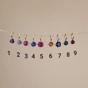 May include: A gold chain featuring nine individual round-cut gemstone charms, each suspended from a small jump ring. The stones are numbered 1 through 9, displaying a variety of colors including purple, light blue, pink, dark blue, and orange-red.