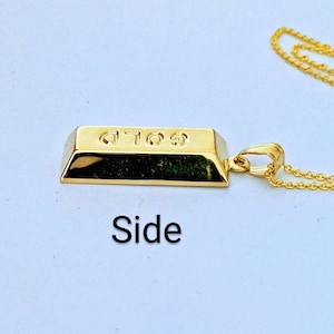 Op de afbeelding: Een goudkleurige hangerketting met een miniatuur goudstaafontwerp. De hanger heeft het woord "GOLD" gegraveerd. De ketting is ook goudkleurig. De afbeelding is van de zijkant genomen.