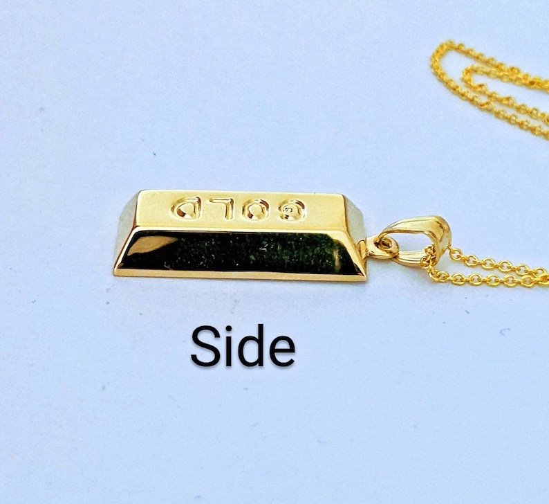 Op de afbeelding: Een goudkleurige hangerketting met een miniatuur goudstaafontwerp. De hanger heeft het woord "GOLD" gegraveerd. De ketting is ook goudkleurig. De afbeelding is van de zijkant genomen.