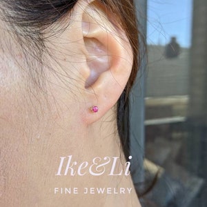 May include: A close-up of a person's ear with a small pink gemstone earring. The earring is set in gold and is a simple, elegant design. The text "Ike & Li Fine Jewelry" is visible in the lower part of the image.