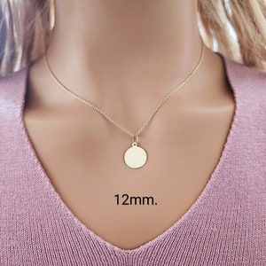 Puede incluir: Un collar de tono dorado que presenta una cadena delicada y un peque&ntilde;o colgante circular liso. El colgante est&aacute; centrado en la cadena y tiene un di&aacute;metro de 12mm. El collar se muestra sobre un fondo de tela de color violeta claro.
