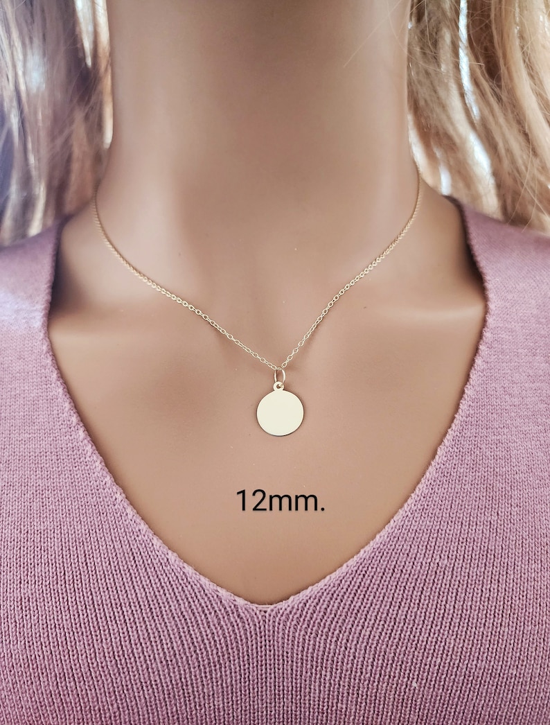 Puede incluir: Un collar de tono dorado que presenta una cadena delicada y un peque&ntilde;o colgante circular liso. El colgante est&aacute; centrado en la cadena y tiene un di&aacute;metro de 12mm. El collar se muestra sobre un fondo de tela de color violeta claro.