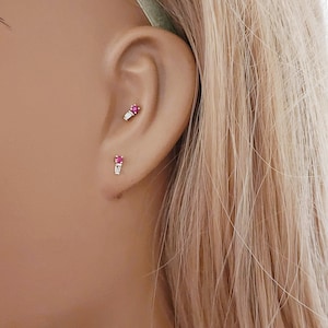 May include: Two gold earrings, each featuring a small round pink gemstone set above a rectangular baguette-cut clear stone. One earring is positioned in the earlobe and the other is placed in the upper cartilage area of the ear.