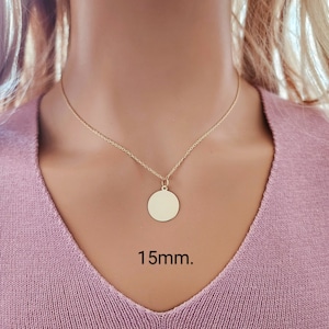 Puede incluir: Un collar de color dorado con una cadena fina y un colgante circular. El colgante es un disco plano y liso de metal dorado con un di&aacute;metro de 15mm. El collar descansa sobre una prenda de tela rosa, mostrando el dise&ntilde;o sencillo de la joya.