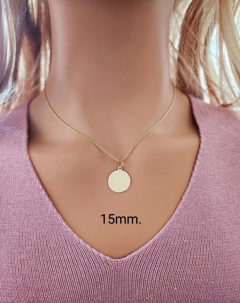 Puede incluir: Un collar de color dorado con una cadena fina y un colgante circular. El colgante es un disco plano y liso de metal dorado con un di&aacute;metro de 15mm. El collar descansa sobre una prenda de tela rosa, mostrando el dise&ntilde;o sencillo de la joya.