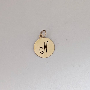 Puede incluir: Un colgante circular de metal dorado con acabado pulido y una argolla en la parte superior. El centro del disco tiene grabada la letra may&uacute;scula N en una fuente cursiva, centrada sobre la superficie plana de la pieza de joyer&iacute;a.