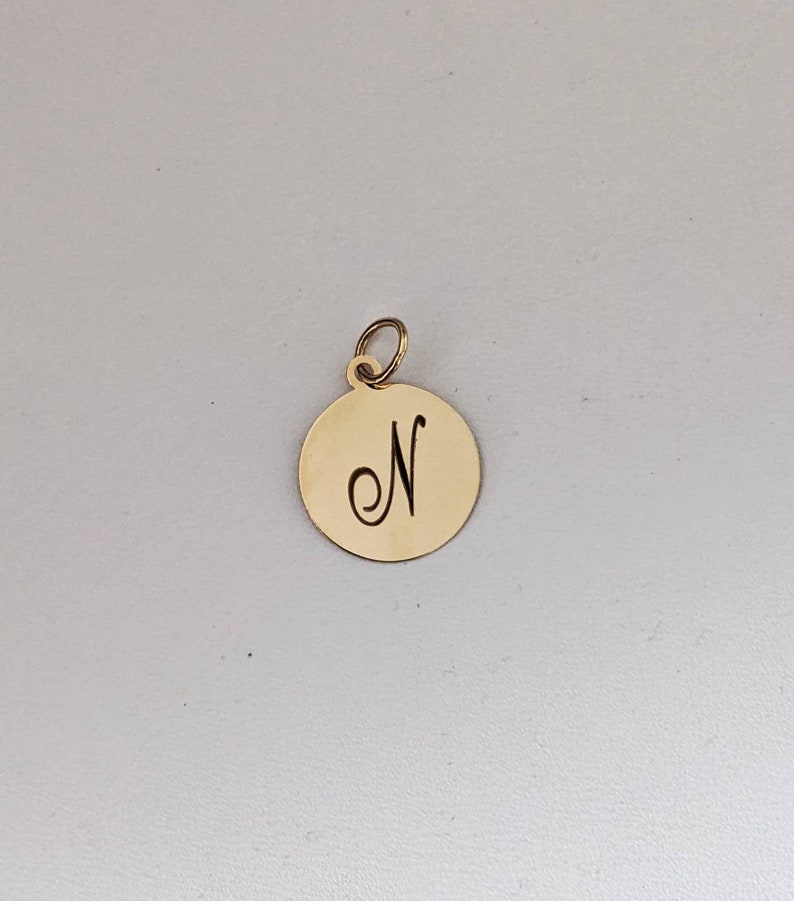 Puede incluir: Un colgante circular de metal dorado con acabado pulido y una argolla en la parte superior. El centro del disco tiene grabada la letra may&uacute;scula N en una fuente cursiva, centrada sobre la superficie plana de la pieza de joyer&iacute;a.