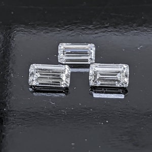May include: Three clear, rectangular, faceted gemstones, likely diamonds, arranged in a triangular pattern on a black surface.