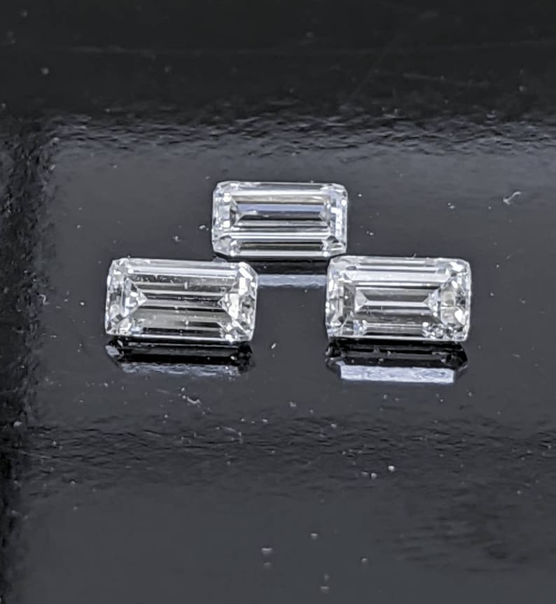 May include: Three clear, rectangular, faceted gemstones, likely diamonds, arranged in a triangular pattern on a black surface.