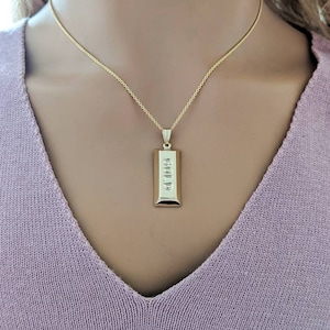 Op de afbeelding: Een goudkleurige ketting met een rechthoekige hanger. De hanger heeft het woord "GOLD" verticaal gegraveerd. De ketting wordt gedragen over een lichtpaarse trui.