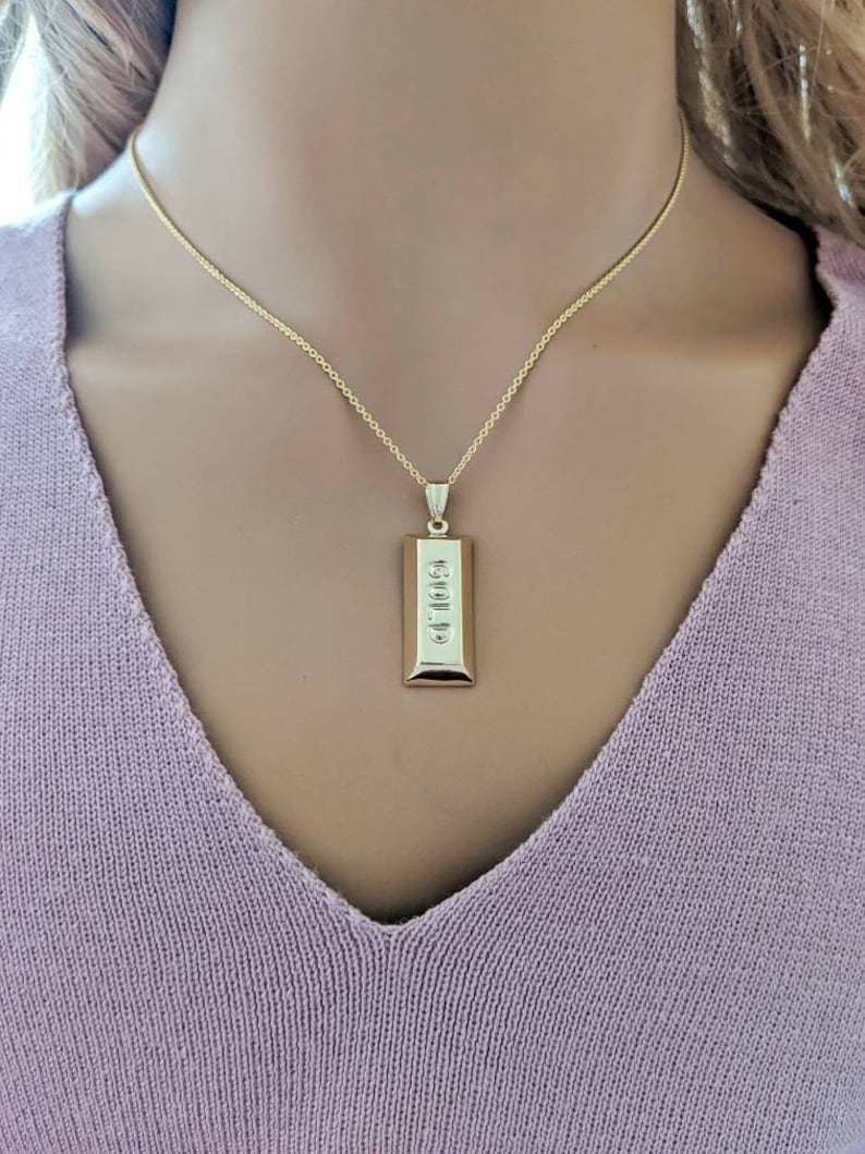 Op de afbeelding: Een goudkleurige ketting met een rechthoekige hanger. De hanger heeft het woord "GOLD" verticaal gegraveerd. De ketting wordt gedragen over een lichtpaarse trui.