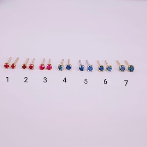 May include: A collection of seven pairs of stud earrings, each with a small, round gemstone. The gemstones are in various colors, including red and blue, set in gold-colored metal. The earrings are numbered 1 through 7.
