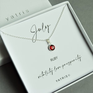 May include: A silver necklace with a red ruby pendant in a white box. The box has the text "July" and "RUBY" printed on it. The text "vitality love prosperity" is also printed on the box.